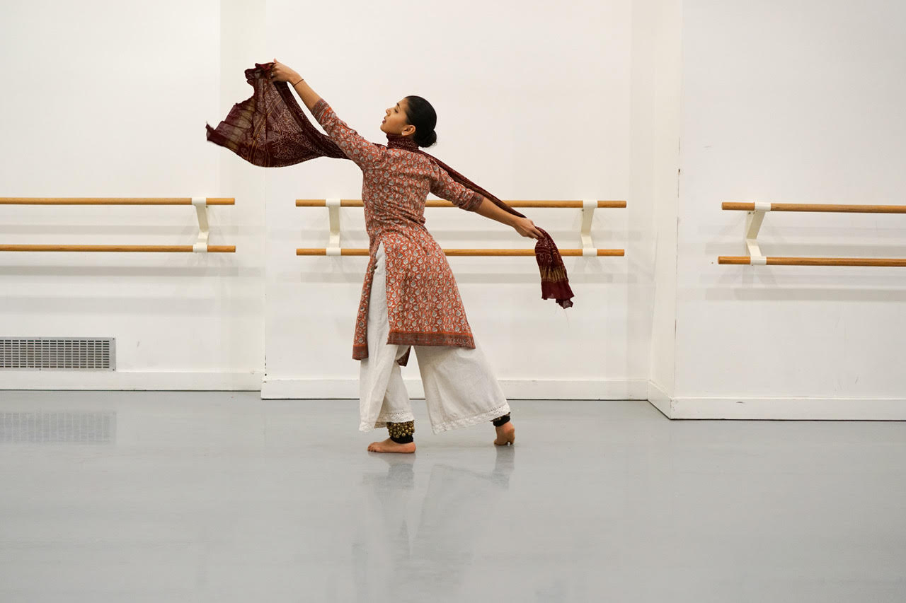 Shared Space Image description: A dancer is in an elongated position with their back towards us. Their arms are extended out and they have one leg extended behind them. They are holding a scarf that traces the top of their shoulder, wearing white pants and a top that has assorted orange and peach shades.