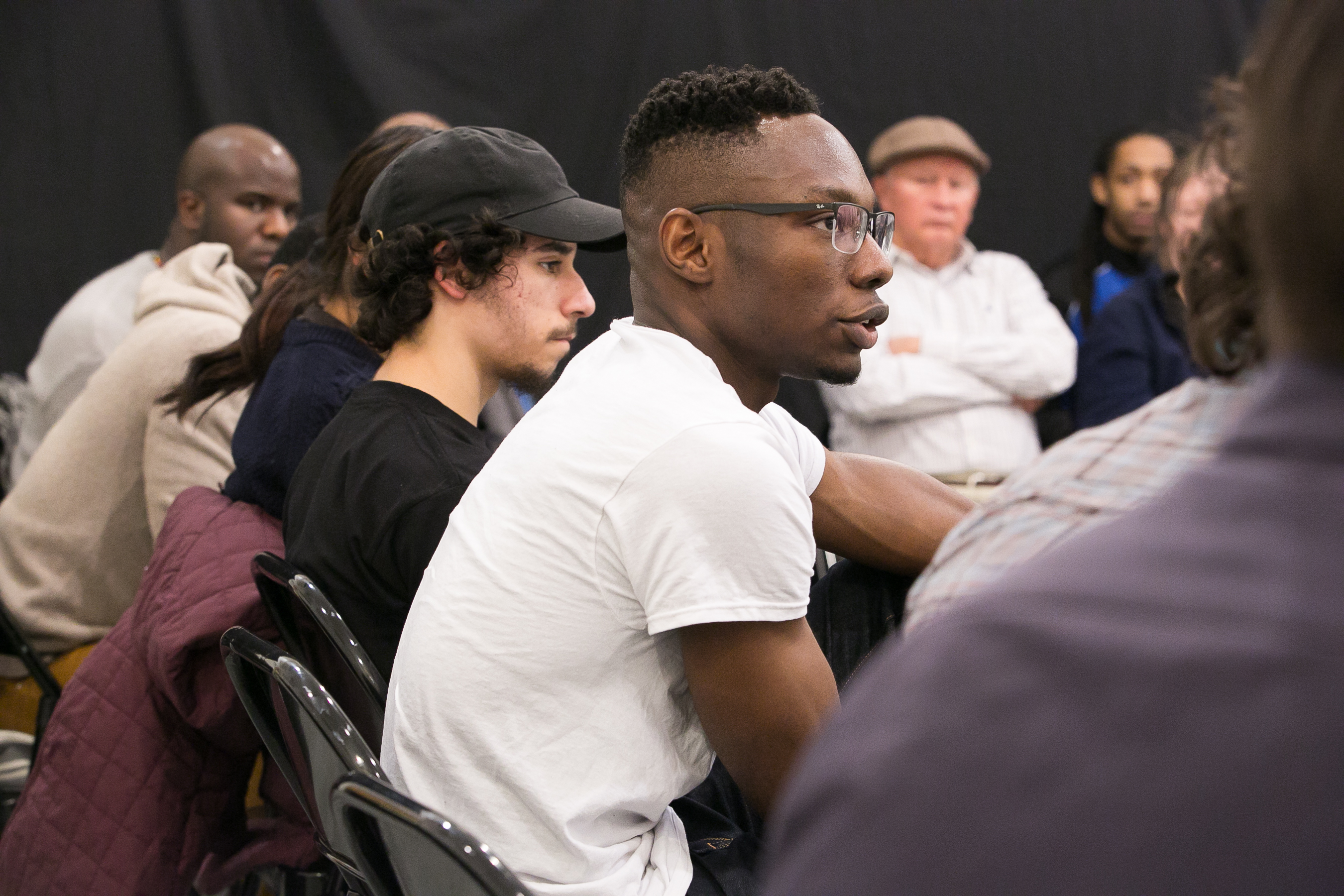 Shared Space Image description: A group of people seated in chairs next to each other. The focus is on a person in a white t-shirt and glasses who is talking.