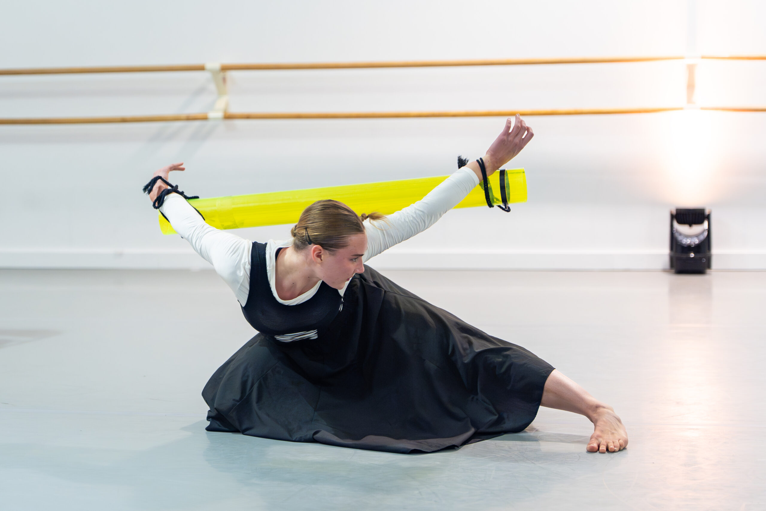 A dancer is on one knee on the ground, and one leg out to the side. The dancer's arms are behind them with each wrist tied to a yellow tube.
