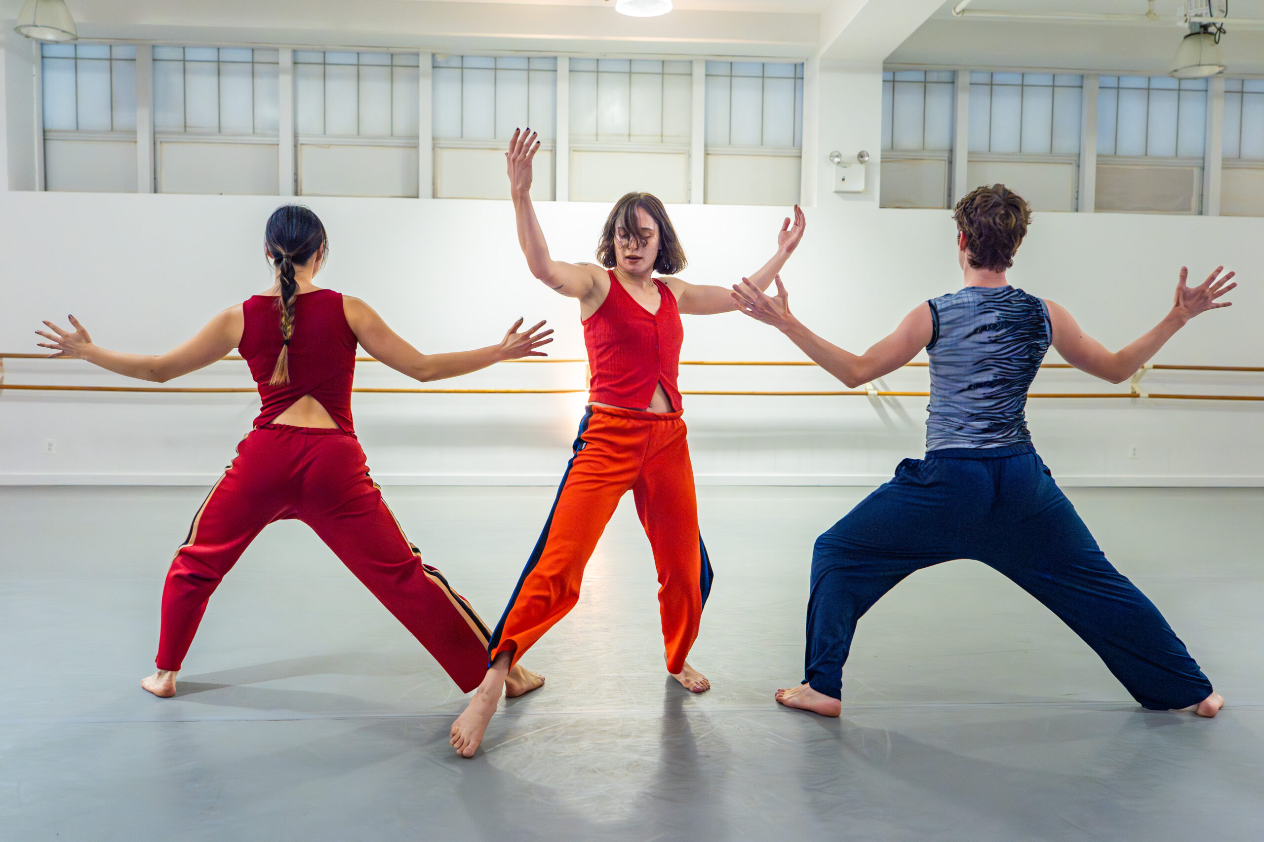 Three dancers stand in a line. Two of the dancers are in a lunge with their backs toward the camera and their arms to the side. The dancer in the middle is facing toward the camera with their arms near their head.