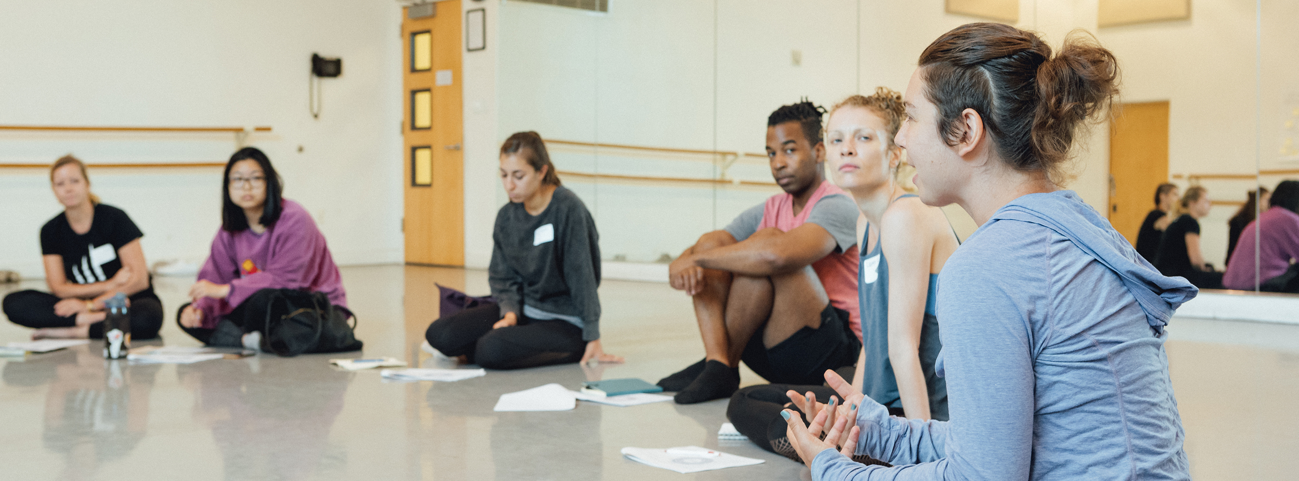 A group of adults are sitting on the floor. The room's focus is on a person with an undercut who is wearing a light blue long sleeve shirt and speaking.