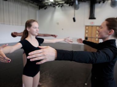 Young dancer in a black leotard and teacher in a black outfit facing one another with their arms out to the side.
