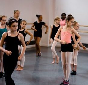 Group of young dancers wearing black and light pink. They have their upper bodies rotated towards the front of the room with the rest of their body facing the side.