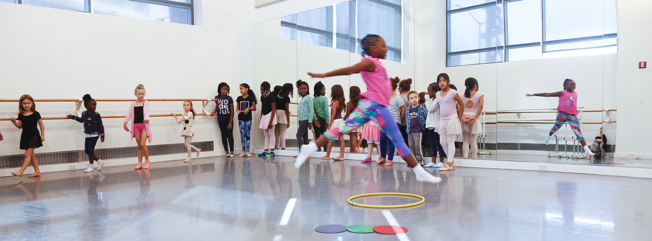 Child leaping over colored dots with a line of children watching her in a dance studio.