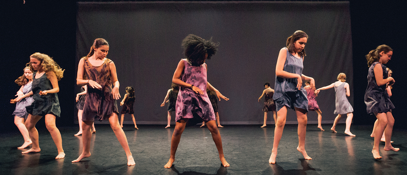The student company is wearing earth tones and are standing in a circle facing outwardly. Their gaze is on their hands which are near their left hip.