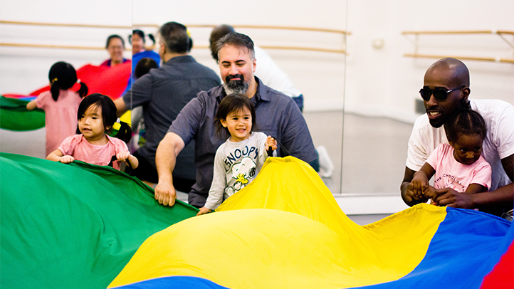 Three toddlers and two parents are holding a green, yellow, blue, and red parachute.