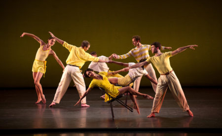 Mark Morris Dance Group in "Dancing Honeymoon." Dressed in all yellow, dancers are in pairs, holding hands and leaning away from one another with their arms extended out. Two of the dancers are sitting and leaning back on black foldable chairs.