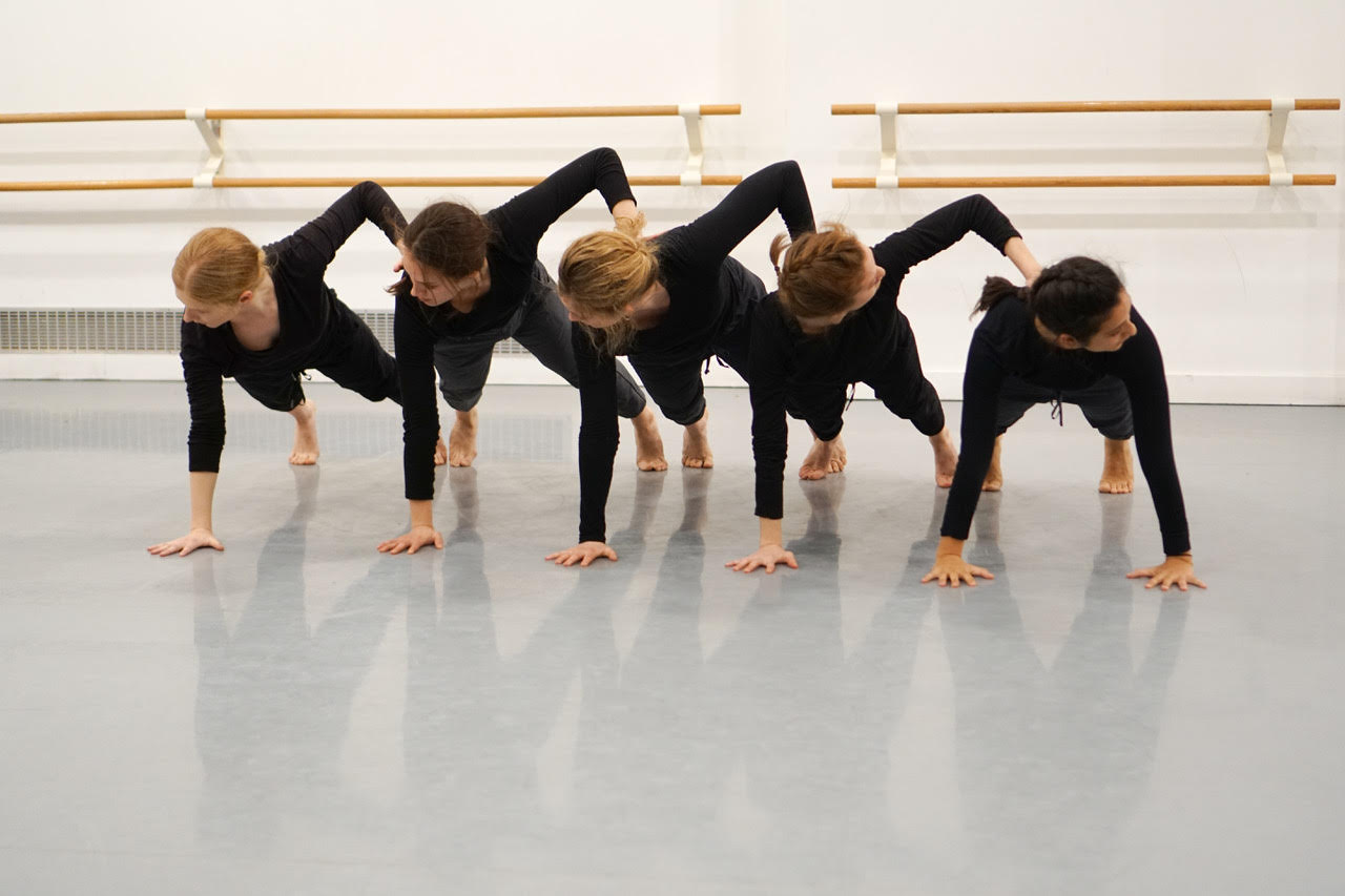 Five dancers are in a pushup position, in a line, next to each other. Their right hand is on the floor and their left hand is on the back of the person to the left of them with their elbows pointed to the sky.