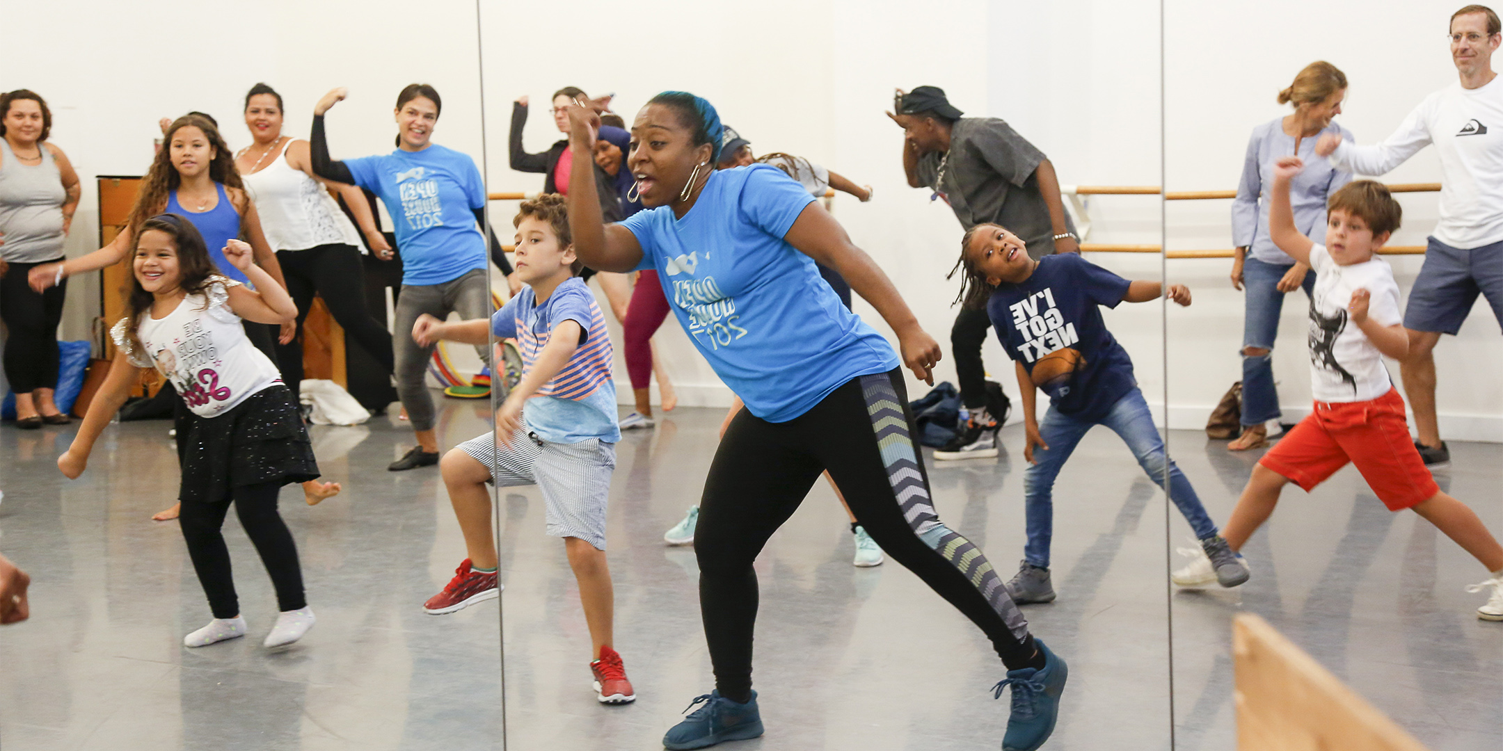 A group of dancers are reflected through a mirror. They are standing and leaning to one side. The dancers are of various ages and are dressed in different assorted colors.