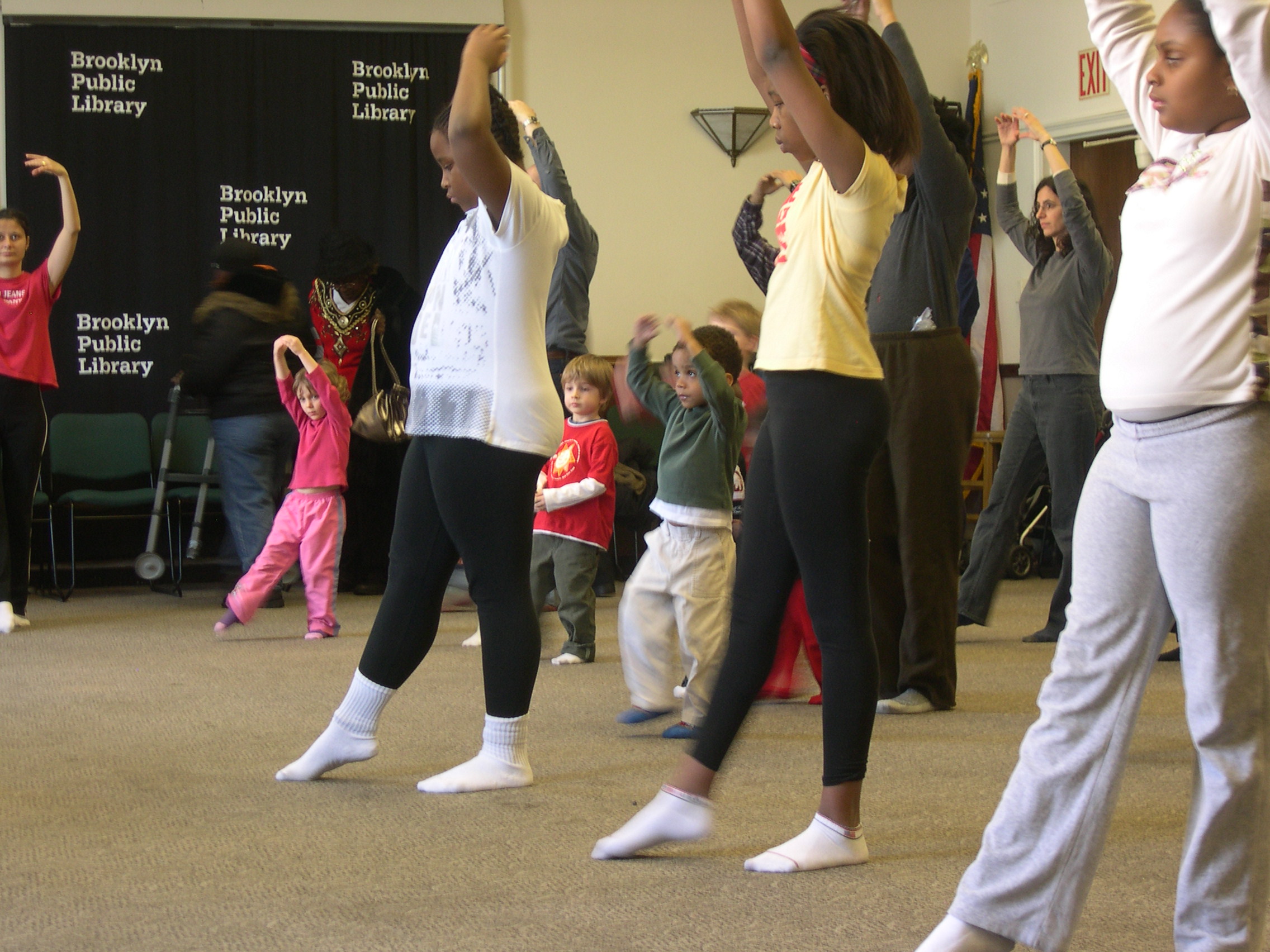 Children of various ages have one foot pointed to the front and are raising their arms creating an 'O' position around their heads.