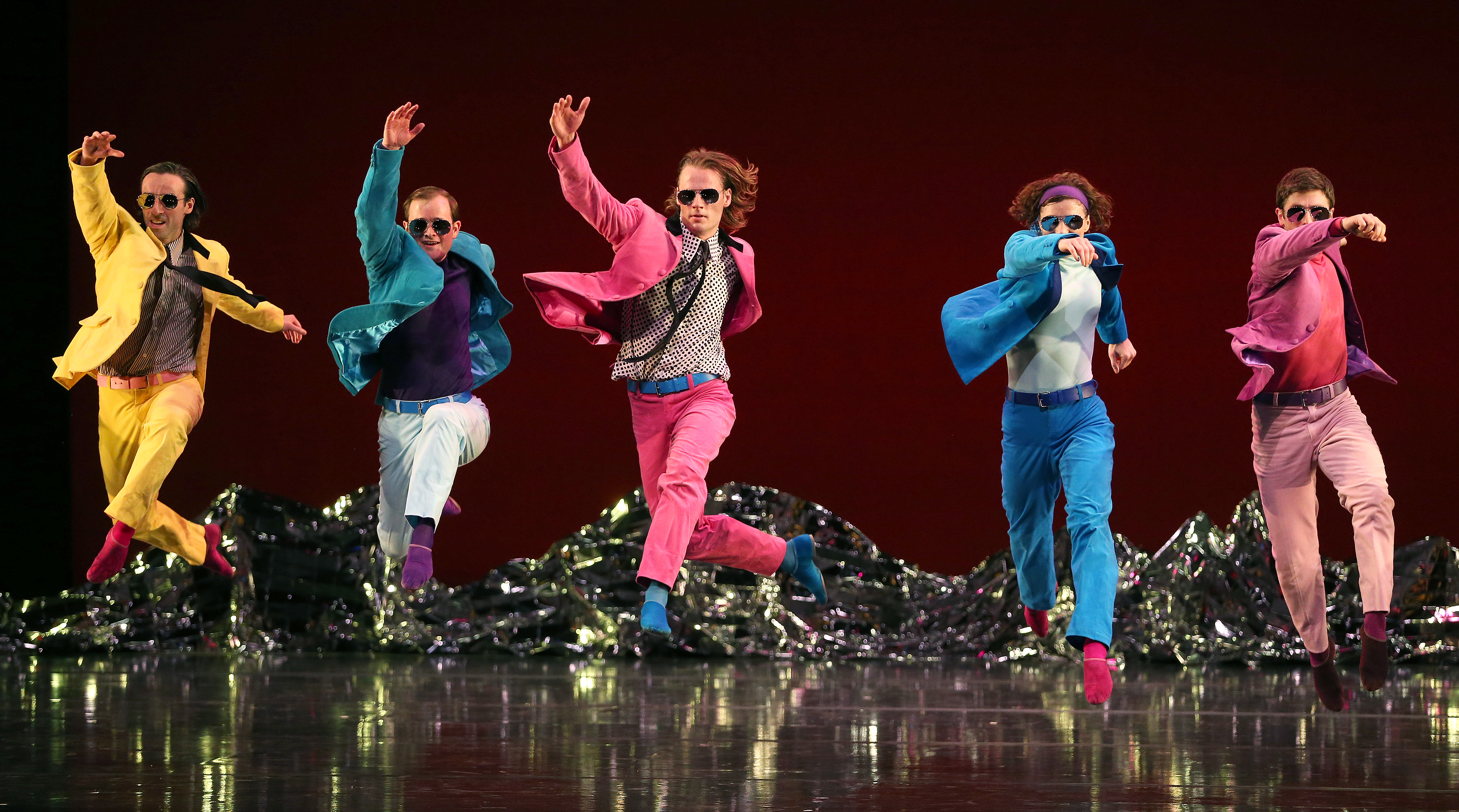 Mark Morris Dance Group in "Pepperland." Five dancers are wearing bright colored suits and sunglasses in mid-air. They are in a running position with their right arm and left leg forward.