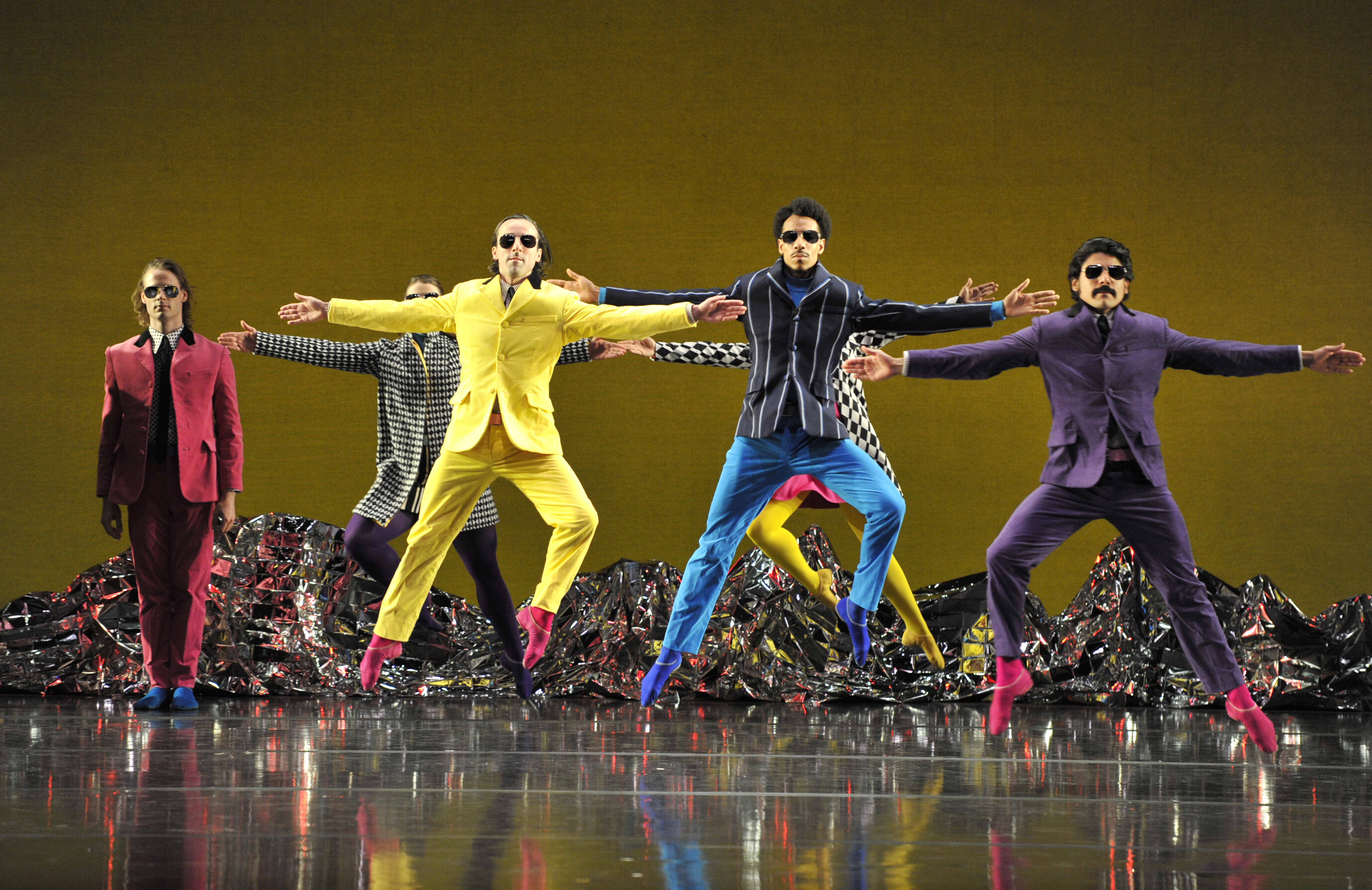 Mark Morris Dance Group in "Pepperland." Wearing an assortment of bright colors, the dancers have their arms out to the side and are in a mid-air position with one leg bent and the other straight.
