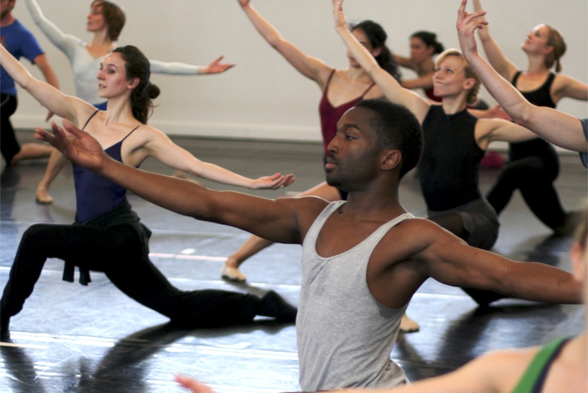Dancers are in a low lung position in dance studio. Their arms are lifted and open, one in front of them and the other behind.