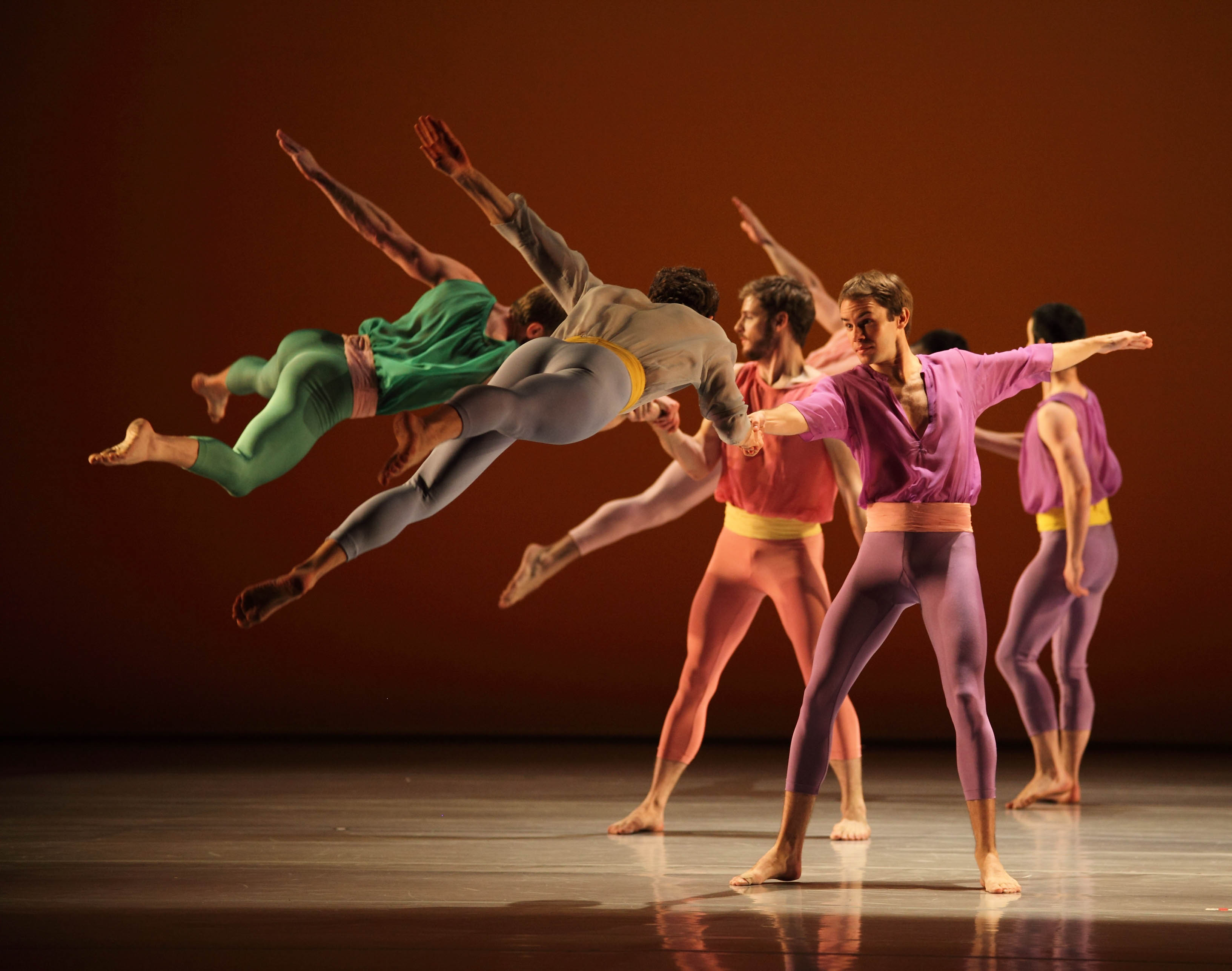Mark Morris Dance Group in "L'Allegro, il Penseroso ed il Moderato." To the left, there are two dancers, one dressed in green and one dressed in grey. They are soaring through the air with their legs behind them and their left arm lifted toward the sky. Their right hand is holding another dancer's hand.