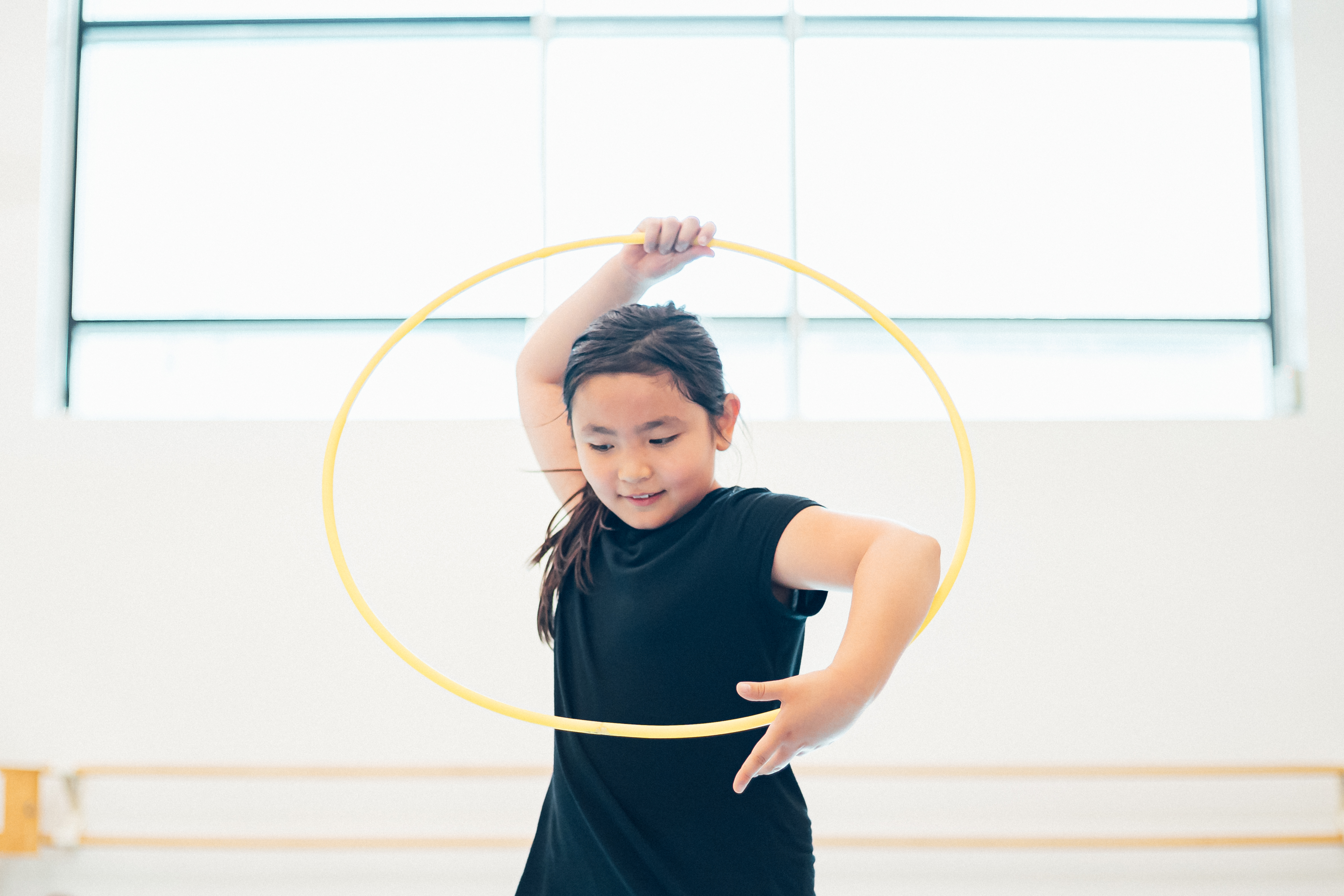 Young dancer holding a yellow hula hoop around their head.