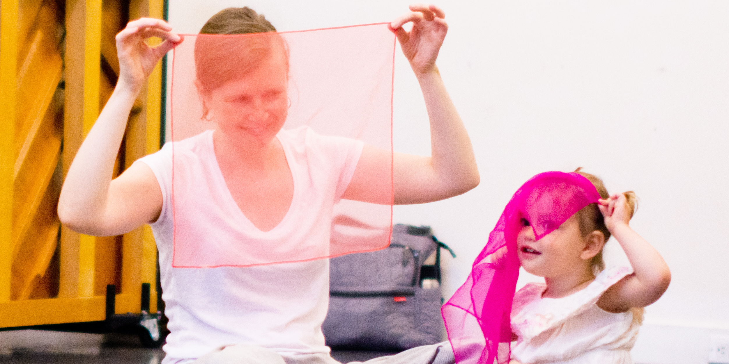Parent sitting on the floor holding a light pink transparent scarf in front of their face. Their child is sitting next to them holding a dark pink transparent scarf on their head, letting it fall in front of their face.