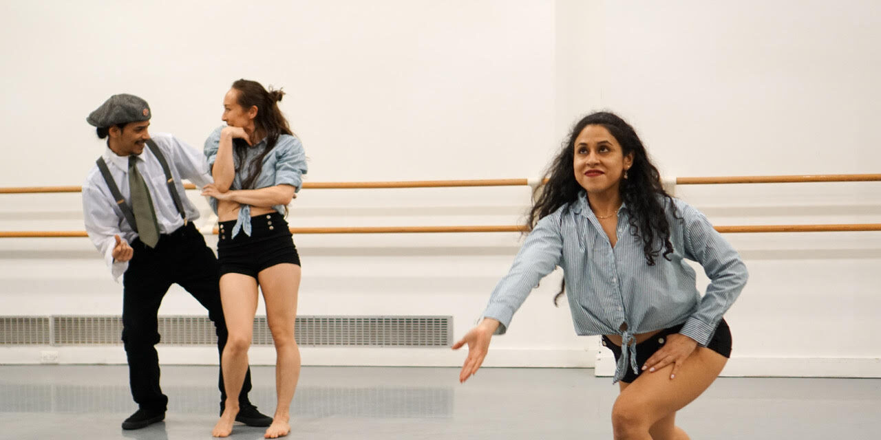 Two dancers are in the corner posed as if they are talking to each other. One dancer is toward the front in a lunged position. Their right arm is lifted forward and their left hand is on their hip.