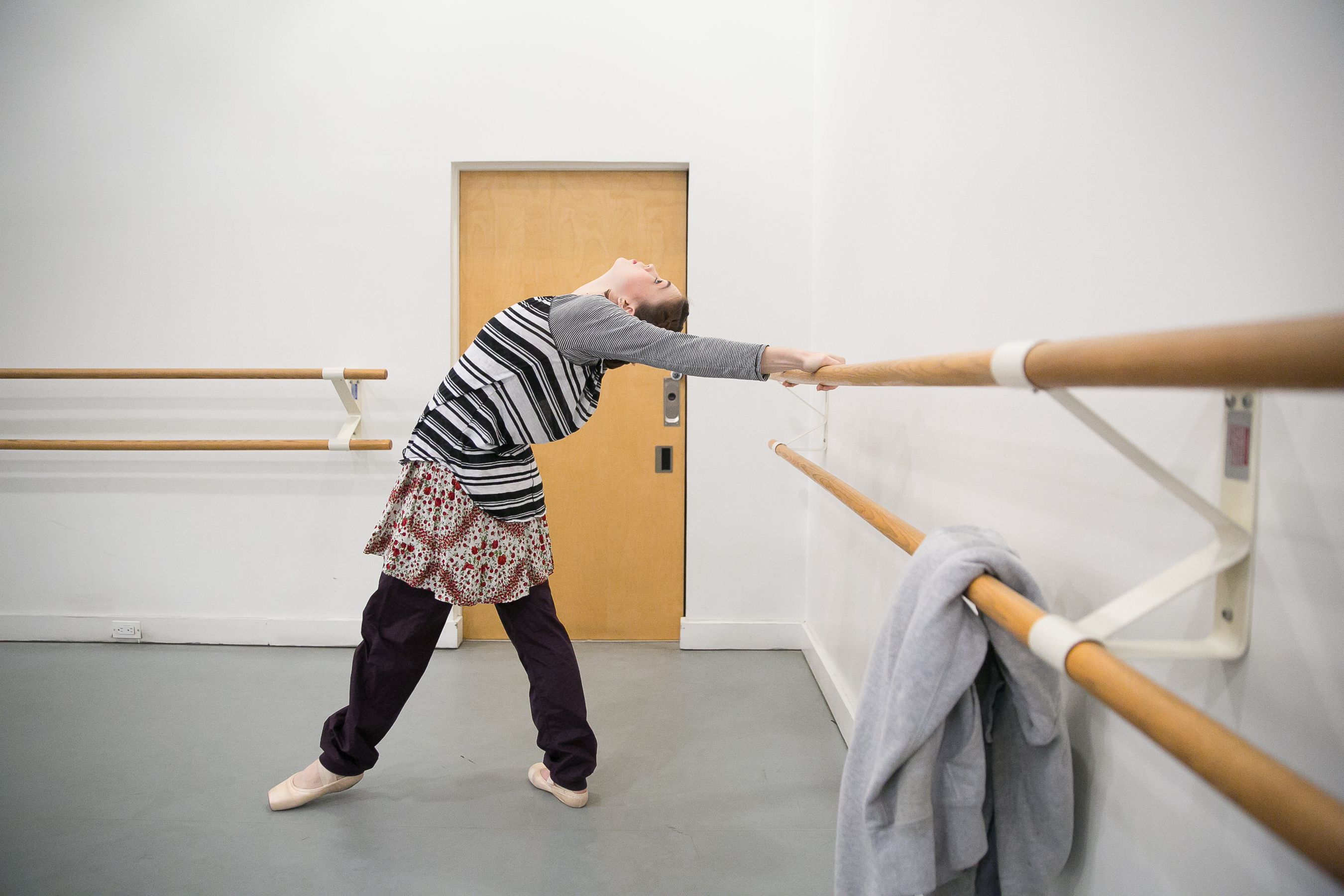 Dancer dressed in a grey striped long sleeve shirt, black pants, pointe shoes, and fabric wrapped around their waist, is arching their back, grabbing the ballet barre behind them.
